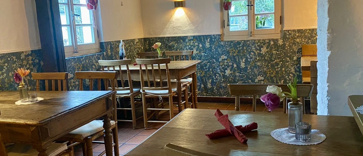 Bistrot - La Souris Rouge Rustikaler Gastraum mit Holztischen, Stühlen, Blumenarrangements und zwei hellen Fenstern.