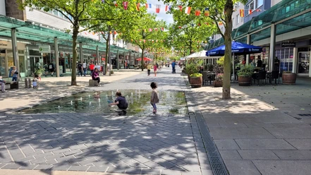 Fußgängerzone 20220615_140547 (c) Tanja Mehl_Erlebnis Bremerhaven.jpg Belebte Fußgängerzone mit Geschäften, Bäumen, bunten Wimpeln und Kindern am Brunnen.