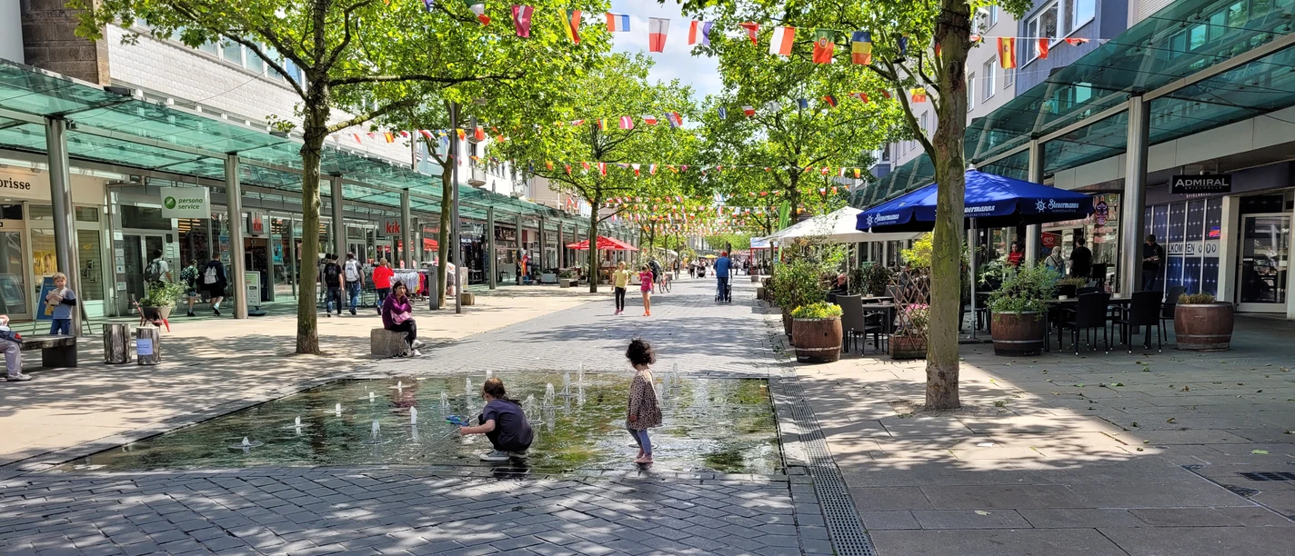 Fußgängerzone 20220615_140547 (c) Tanja Mehl_Erlebnis Bremerhaven.jpg Belebte Fußgängerzone mit Geschäften, Bäumen, bunten Wimpeln und Kindern am Brunnen.