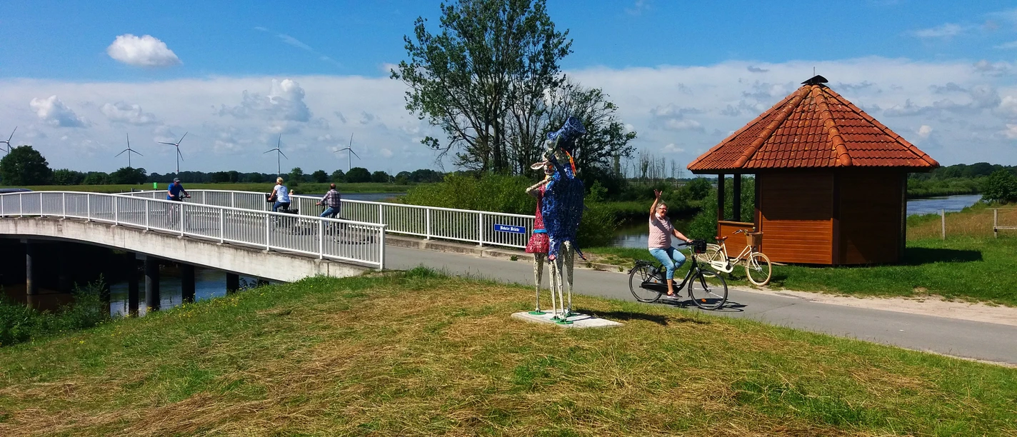 Lieblingsorte-Bokeler Brücke-Vreschen Bokel-Apen-Foto Sandra Hinrichs.jpg Radfahrer queren die Bokeler Brücke nahe einer bunten Skulptur und Holzhütte.