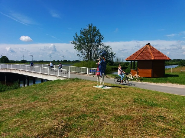 Lieblingsorte-Bokeler Brücke-Vreschen Bokel-Apen-Foto Sandra Hinrichs.jpg Radfahrer queren die Bokeler Brücke nahe einer bunten Skulptur und Holzhütte.