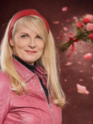Barbara Ruscher Lächelnde Frau mit blonden Haaren, rotem Haarband und pinker Jacke vor Rosen und Blütenblättern.