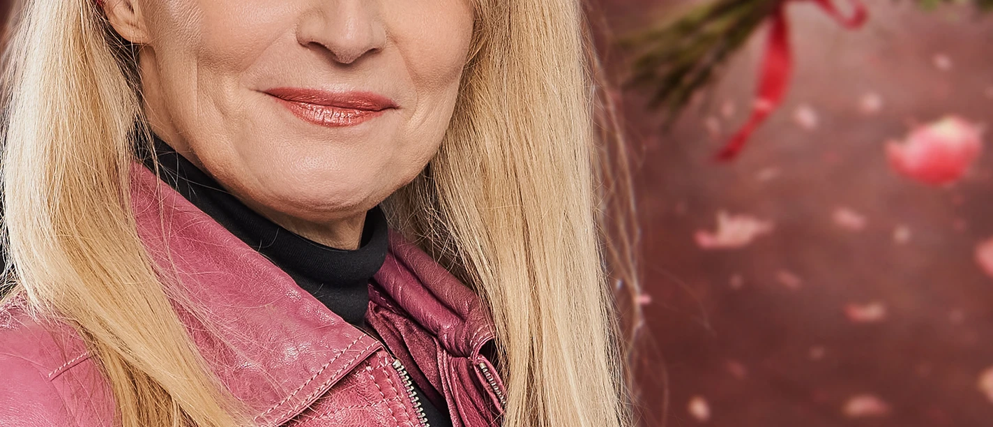 Barbara Ruscher Lächelnde Frau mit blonden Haaren, rotem Haarband und pinker Jacke vor Rosen und Blütenblättern.