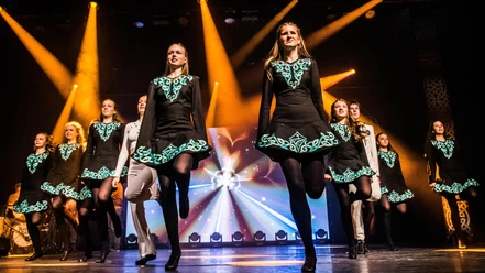 The Arc of Irish Dance.jpg Irish-Dance-Gruppe in schwarzen Kostümen tanzt auf beleuchteter Bühne.