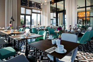 Helles Restaurant mit gedeckten Holztischen, türkisen Stühlen und geometrischem Fliesenboden.