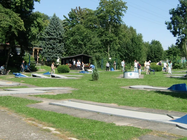 Minigolfplatz_Gebhardshagen_HD_16zu9.jpg Gäste spielen bei sonnigem Wetter auf einer grünen Minigolfanlage mit grauen Bahnen.