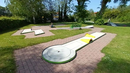 Minigolfanlage_Lebenstedt_HD_16zu9.jpg Mehrere helle Minigolfbahnen mit Hindernissen auf grünem Rasen zwischen Bäumen.