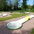 Minigolfanlage_Lebenstedt_HD_16zu9.jpg Mehrere helle Minigolfbahnen mit Hindernissen auf grünem Rasen zwischen Bäumen.