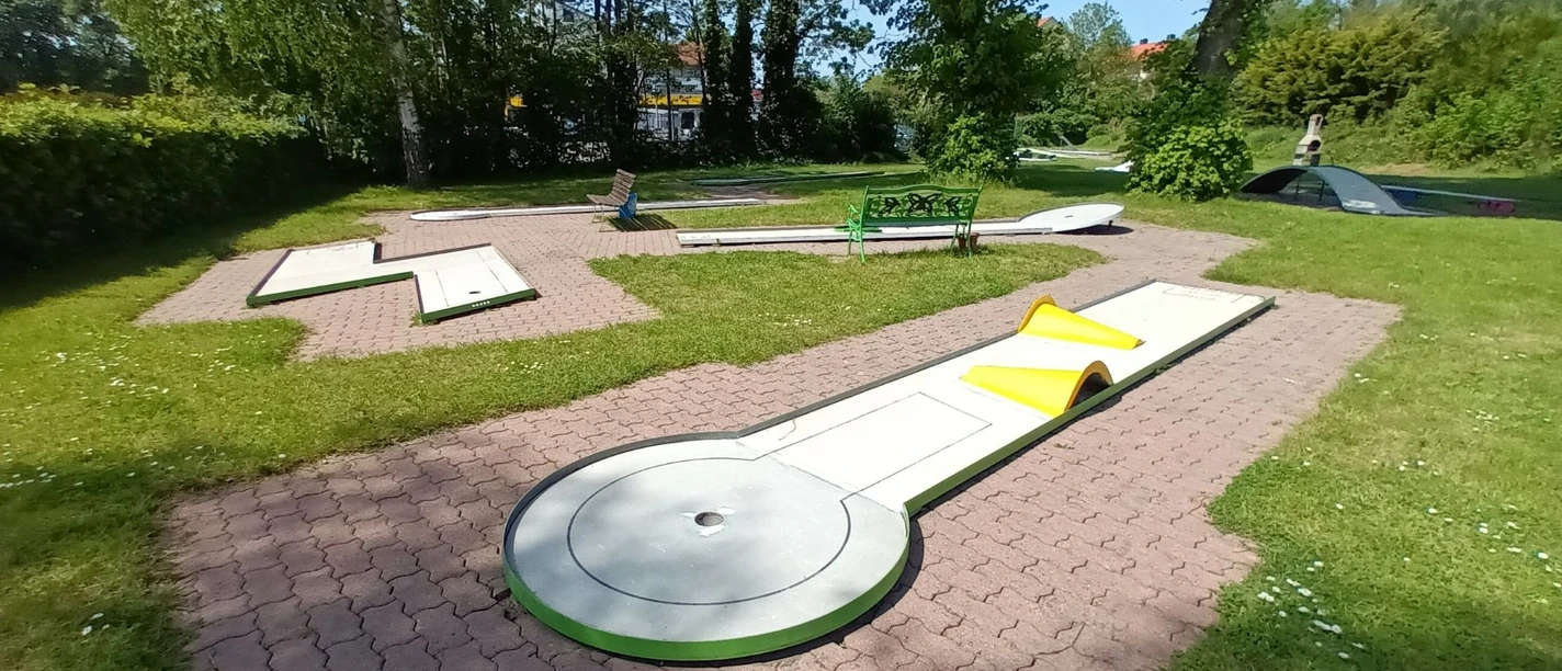Minigolfanlage_Lebenstedt_HD_16zu9.jpg Mehrere helle Minigolfbahnen mit Hindernissen auf grünem Rasen zwischen Bäumen.