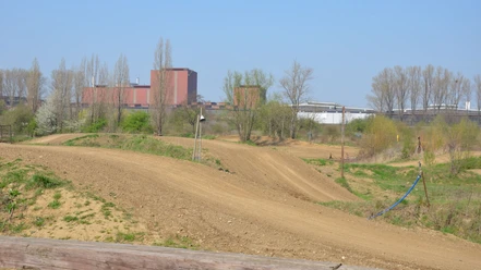 Motocross_Salzgitter(3)_HD_16zu9.JPG Sandige Motocross-Strecke mit Hügeln vor roten Industriegebäuden und blauem Himmel.