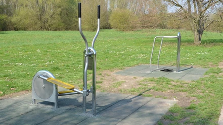 Freisportfläche_Salzgittersee_Outdoorfitnessgeräte_16zu9.JPG Zwei Outdoor-Fitnessgeräte aus Metall auf Schutzmatten im grünen Park.
