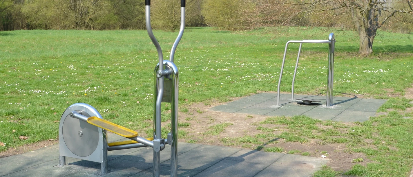 Freisportfläche_Salzgittersee_Outdoorfitnessgeräte_16zu9.JPG Zwei Outdoor-Fitnessgeräte aus Metall auf Schutzmatten im grünen Park.