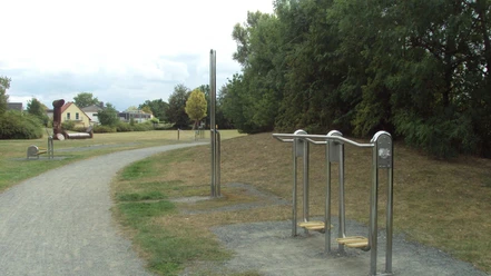 Fitnesspark_Thiede_HD_16zu9.JPG Mehrere Outdoor-Fitnessgeräte aus Metall stehen entlang eines Kieswegs in einer Parkanlage.
