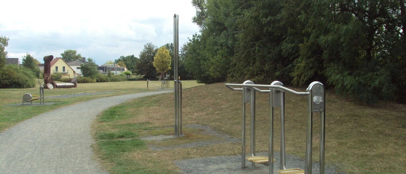 Fitnesspark_Thiede_HD_16zu9.JPG Mehrere Outdoor-Fitnessgeräte aus Metall stehen entlang eines Kieswegs in einer Parkanlage.