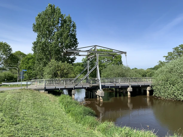 alte Staaßenbrücke_Antje Geisler.jpg Alte graue Klappbrücke über einem Fluss, umgeben von grünen Bäumen unter blauem Himmel.