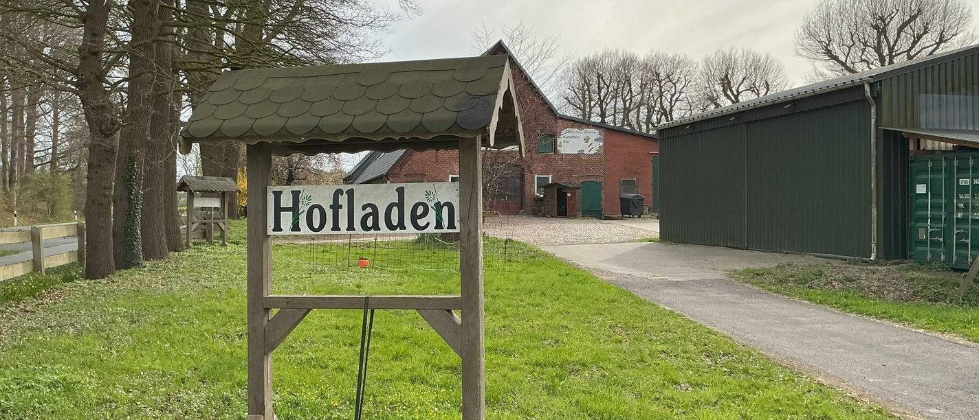 Hofladen in Stendorf Holzschild mit der Aufschrift Hofladen an einer Hofeinfahrt vor einem roten Backsteingebäude.