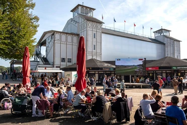 Galopprennbahn Köln-Weidenpesch Menschen sitzen vor der Tribüne der Rennbahn Weidenpesch, umgeben von Ständen und großem Bildschirm.