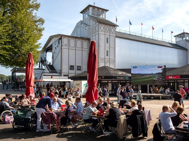 Galopprennbahn Köln-Weidenpesch Menschen sitzen vor der Tribüne der Rennbahn Weidenpesch, umgeben von Ständen und großem Bildschirm.