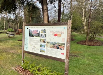 Infotafel zum "Kleinen Haus im Moor" Infotafel vor einem grünen Spielplatz mit kleinem roten Holzhaus, Sandkasten und Bäumen.