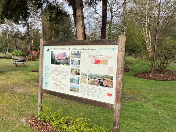 Infotafel zum "Kleinen Haus im Moor" Infotafel vor einem grünen Spielplatz mit kleinem roten Holzhaus, Sandkasten und Bäumen.