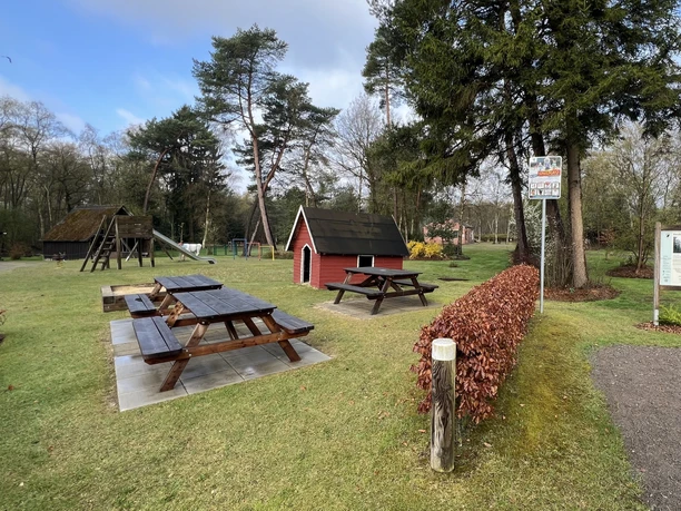 Rastplatz am Kleinen Haus im Moor Spielplatz mit Picknicktischen, rotem Spielhaus und Infotafel auf einer grünen Wiese vor Bäumen.