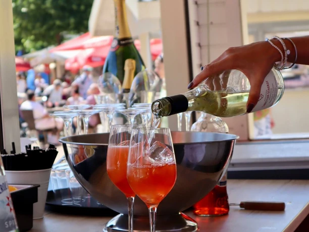 Fisch- und Genussmarkt auf der Rennbahn Köln Weidenpesch Hand schenkt Wein in zwei Aperolgläser an einem offenen Marktstand mit Menschen im Hintergrund