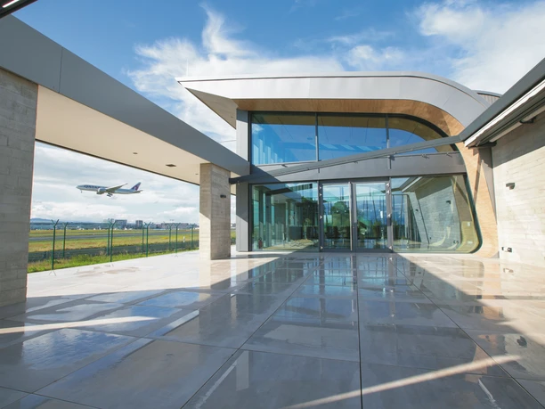 FRALounge Frankfurt Airport - Außenansicht einer modernen Flughafen-Lounge Außenansicht einer modernen Flughafen-Lounge mit großer Glasfront und Terrasse, im Hintergrund ein startendes Flugzeug vor blauem Himmel.