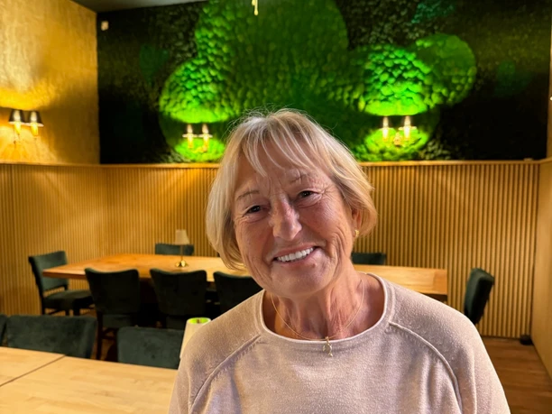 Grandma Anelie Older woman smiling in warmly lit restaurant in front of green wall decoration.
