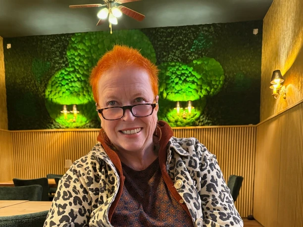 Grandma Gitta Person with short red hair sitting smiling on chair in warmly lit restaurant