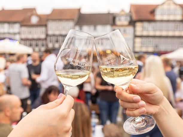 Weinmarkt Celle - Genuss pur vor historischer Fachwerkkulisse Zwei Personen stoßen mit Gläsern Weißwein auf dem Celler Weinmarkt auf dem Großen Plan an, umgeben von fröhlichen Besuchern und den historischen Fachwerkhäusern der Celler Altstadt.