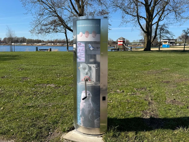 Souvenirmedaillen-Automat am Ankerplatz Ein Souvenirmedaillen-Automat mit Otter-Motiv lädt auf grüner Wiese am Wasser zum Kurbeln ein.
