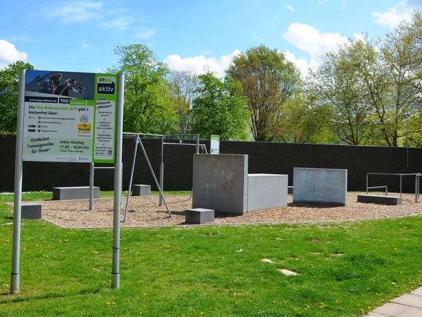 Parcours_Salzgitter(1)_HD_16zu9.JPG Ein Fitness-Parcours im Freien mit Infoschild, Betonblöcken und Metallstangen auf einer Wiese.