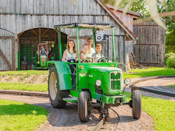 treggerbahn.jpg Familie fährt lachend in einem grünen Traktor auf Schienen vor einer alten Holzscheune.