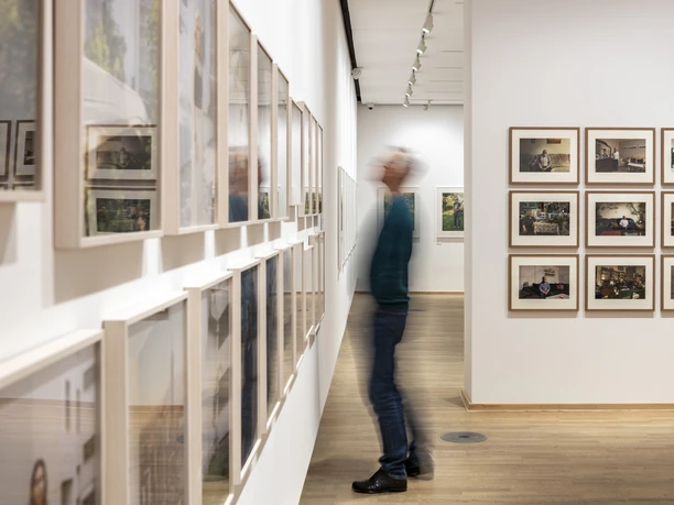 Die Photographische Sammlung/SK Stiftung Kultur Ausstellungsraum mit vielen gerahmten Fotografien und einer verschwommenen Person beim Betrachten