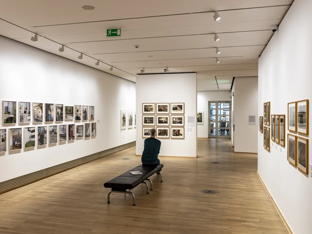 The Photographic Collection/SK Stiftung Kultur Bright gallery with photographs, person sitting on a bench and looking at a picture wall.