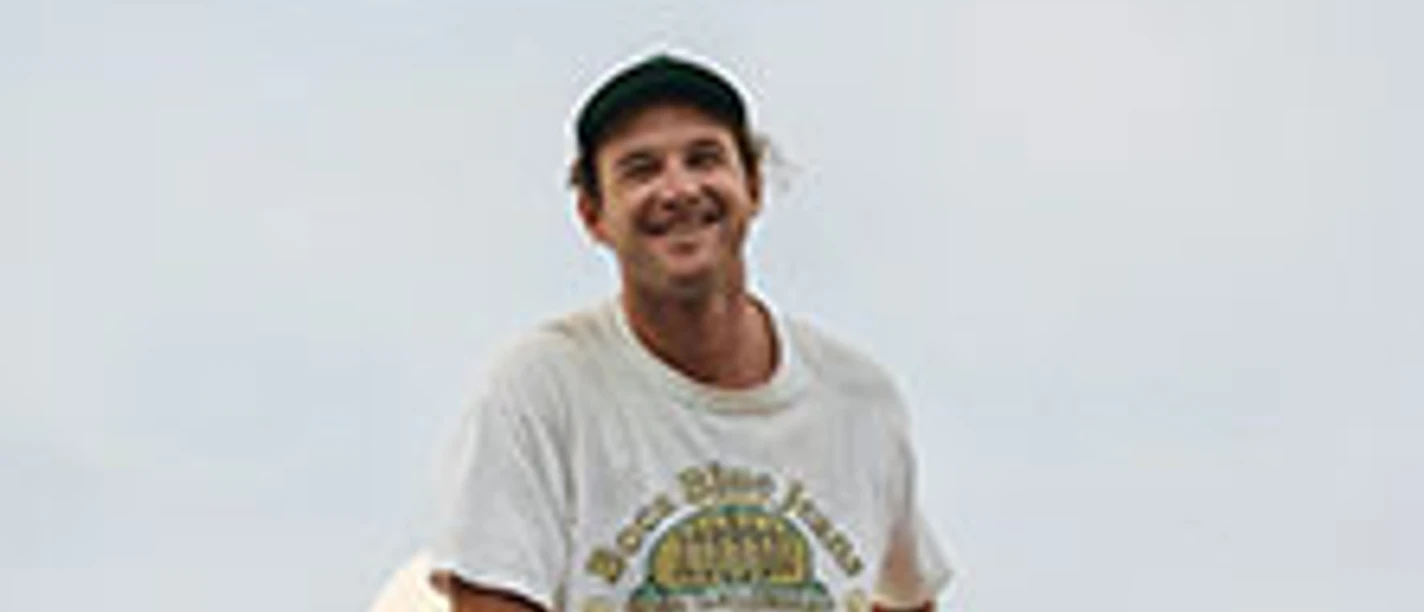 Ein lächelnder Mann sitzt entspannt im Freien vor hellem Himmel und trägt T-Shirt und Kappe.
