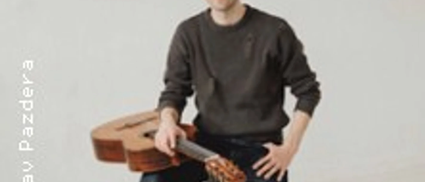 Ein Mann sitzt auf einem Hocker in hellem Raum und hält eine Akustikgitarre neben sich.