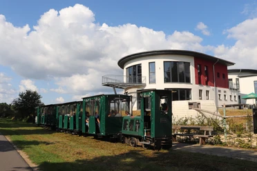 Ströher Moorbahn.JPG Grüne Moorbahn mit Fahrgästen vor einem modernen Gebäude bei blauem Himmel.