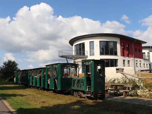 Ströher Moorbahn.JPG Grüne Moorbahn mit Fahrgästen vor einem modernen Gebäude bei blauem Himmel.