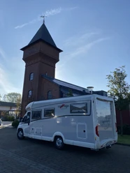 Wohnmobil am Eisenhüttenturm.jpg Ein helles Wohnmobil parkt direkt vor einem markanten Backsteinturm mit spitzem Dach bei blauem Himmel.