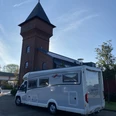 Wohnmobil am Eisenhüttenturm.jpg Ein helles Wohnmobil parkt direkt vor einem markanten Backsteinturm mit spitzem Dach bei blauem Himmel.