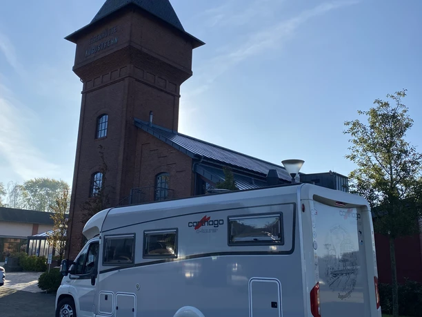 Wohnmobil am Eisenhüttenturm.jpg Ein helles Wohnmobil parkt direkt vor einem markanten Backsteinturm mit spitzem Dach bei blauem Himmel.