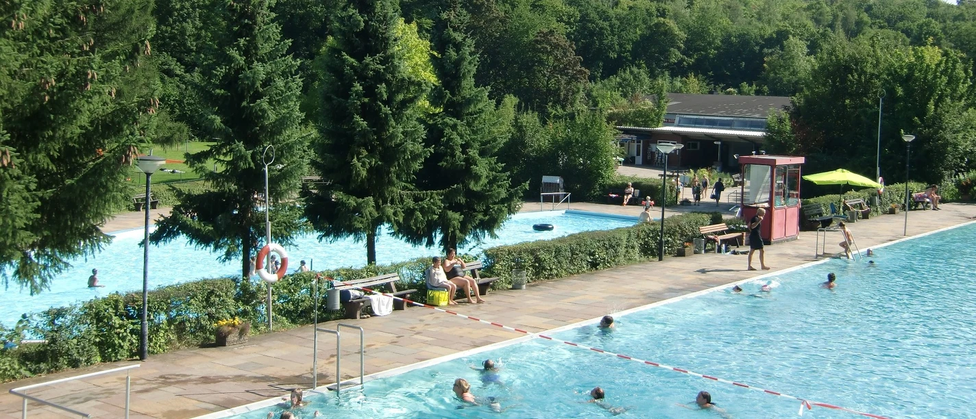 Waldschwimmbad_Salzgitter-Gebhardshagen_HD_16zu9.jpg Freibad am Waldrand mit schwimmenden Gästen in zwei blauen Becken und großen Nadelbäumen.