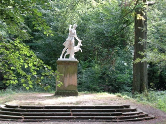 Schlosspark Dittersbach Skulptur der Diana Figur mit einem Hirsch auf einem Sockel, umgeben von Bäumen und Natur.