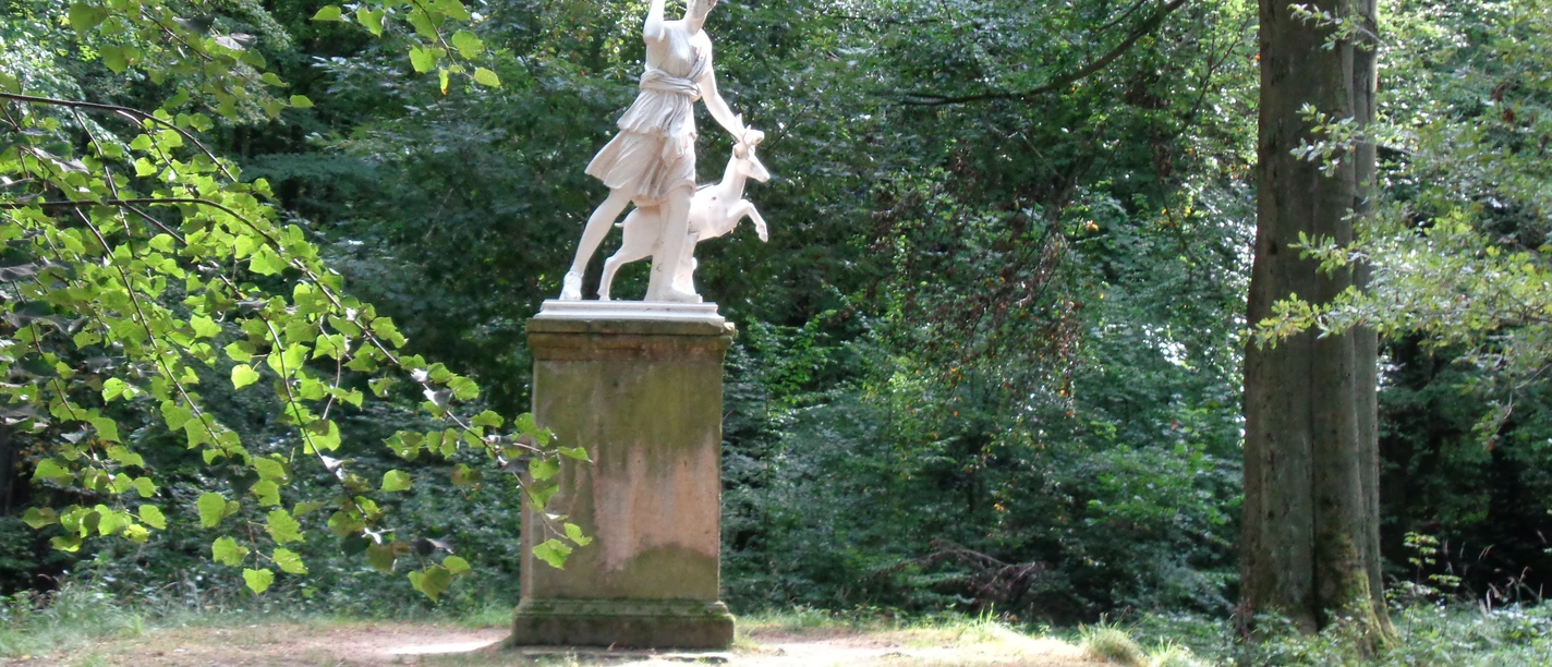 Schlosspark Dittersbach Skulptur der Diana Figur mit einem Hirsch auf einem Sockel, umgeben von Bäumen und Natur.