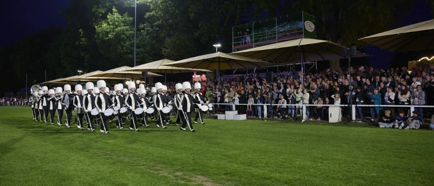 Advendo Sneek beim Finale (Mittel) Eine uniformierte Marching Band marschiert nachts bei Flutlicht auf Rasen vor einem Publikum.