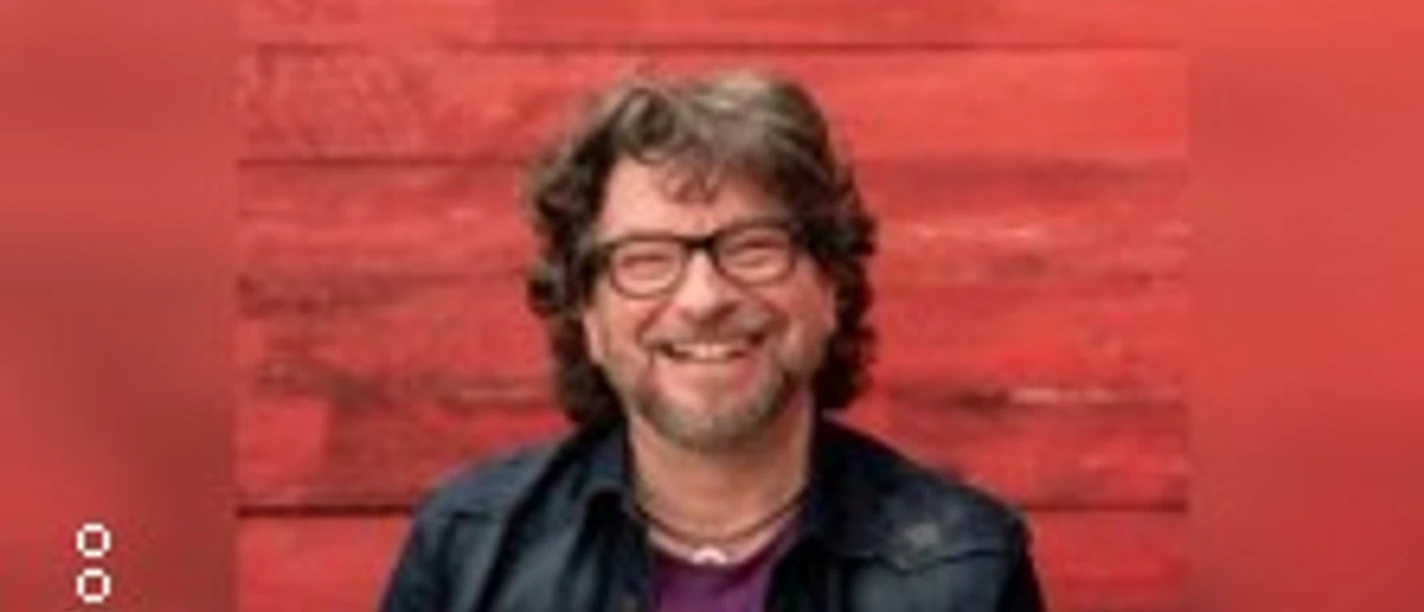 Eine Person mit Brille sitzt lächelnd vor einer roten Holzwand.