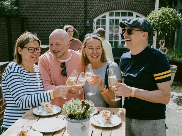 Vier Erwachsene stoßen lachend mit Sektgläsern an. Auf dem Tisch vor ihnen stehen kleine Häppchen.