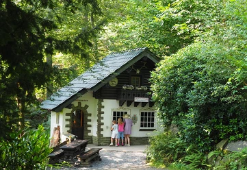 Rotkäppchen Kleines Märchenhaus im Wald, vor dem drei Personen am Fenster stehen.