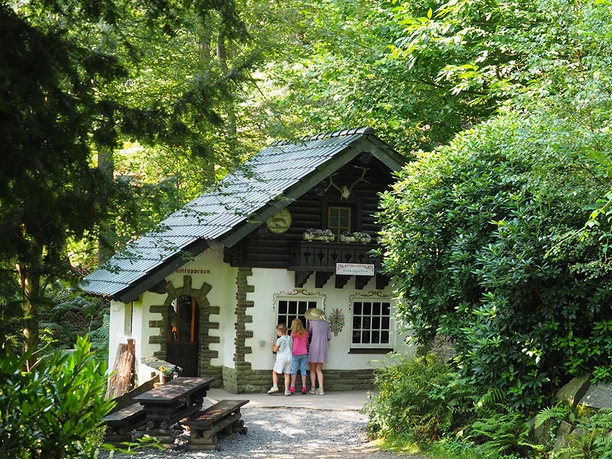 Rotkäppchen Kleines Märchenhaus im Wald, vor dem drei Personen am Fenster stehen.